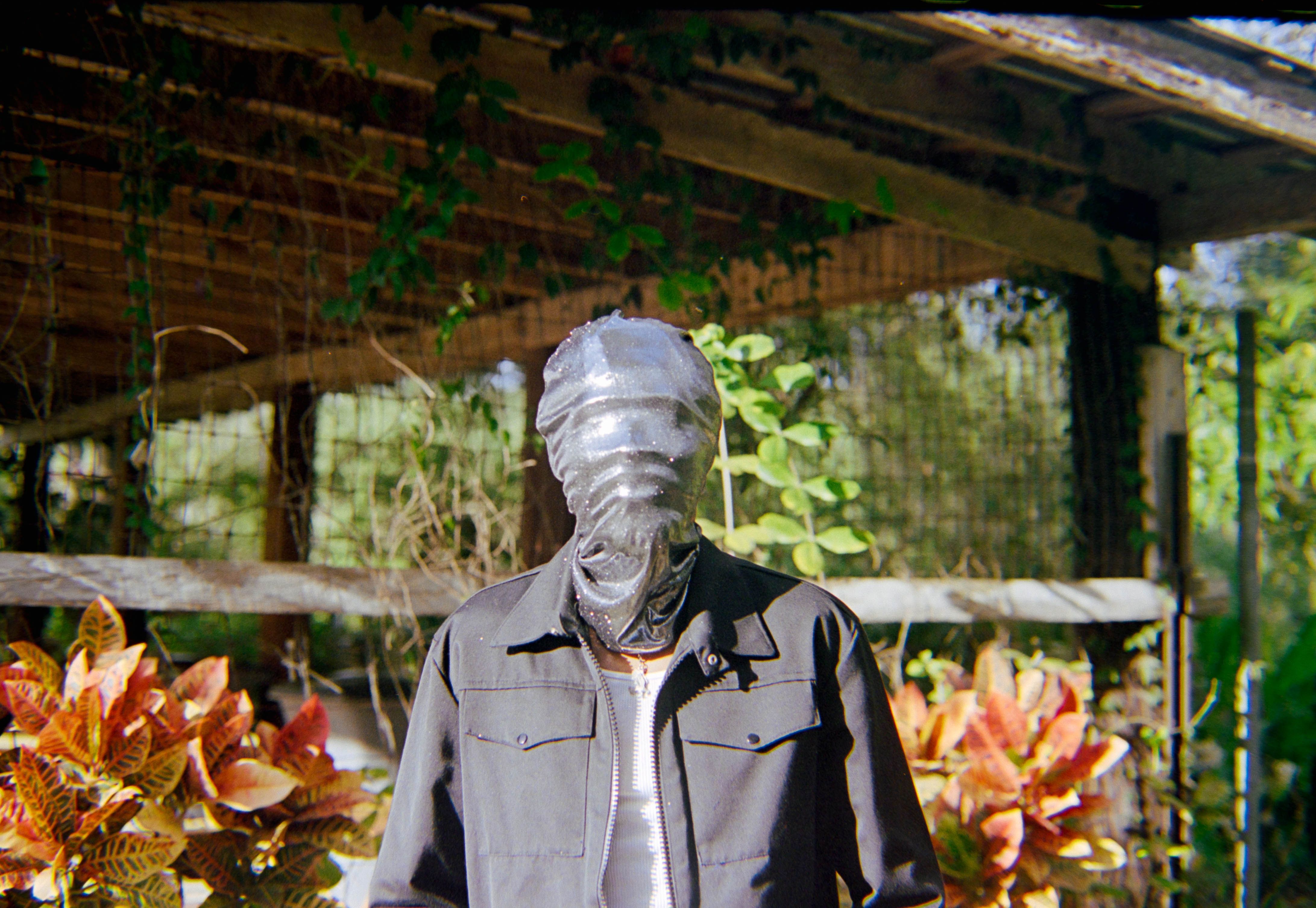 A person with a veiled face stands outdoors amidst vibrant foliage, creating a sense of mystery.