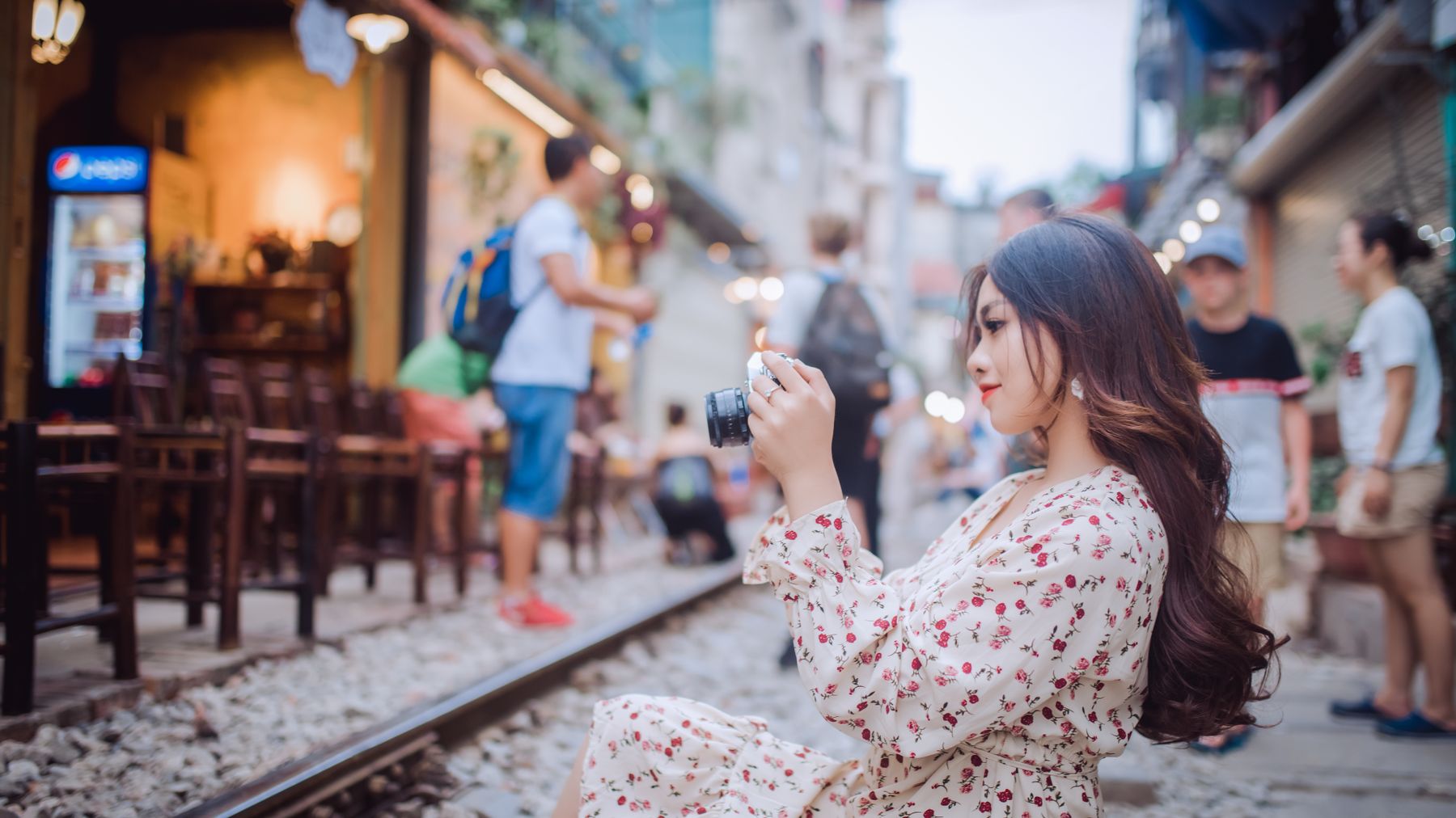 Woman in floral dress taking a photo on a vibrant street with railway.