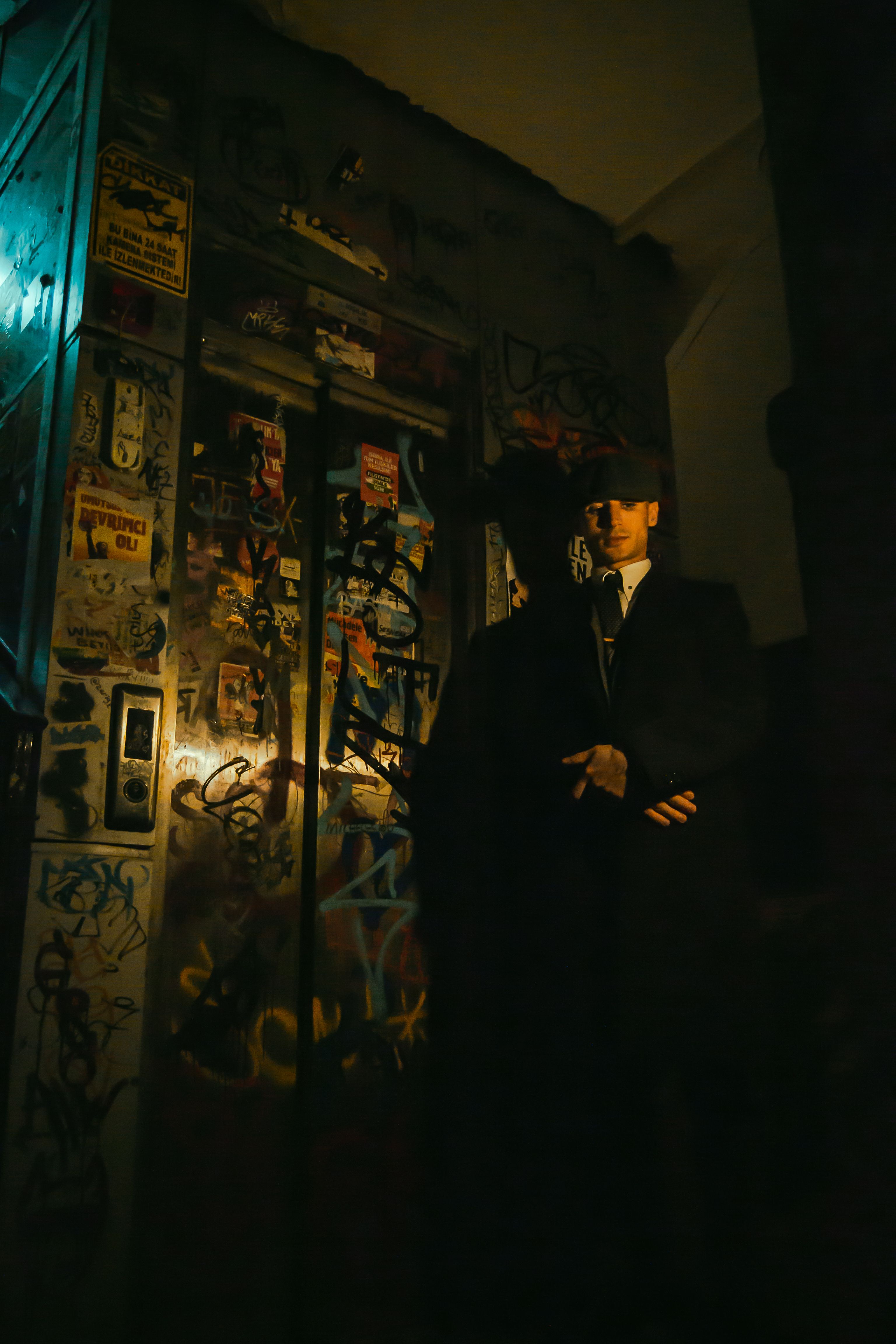 A man in a suit stands near a graffiti-covered wall under moody lighting.