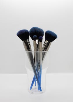 Set of elegant makeup brushes with blue bristles in a clear glass cup, ideal for beauty enthusiasts.