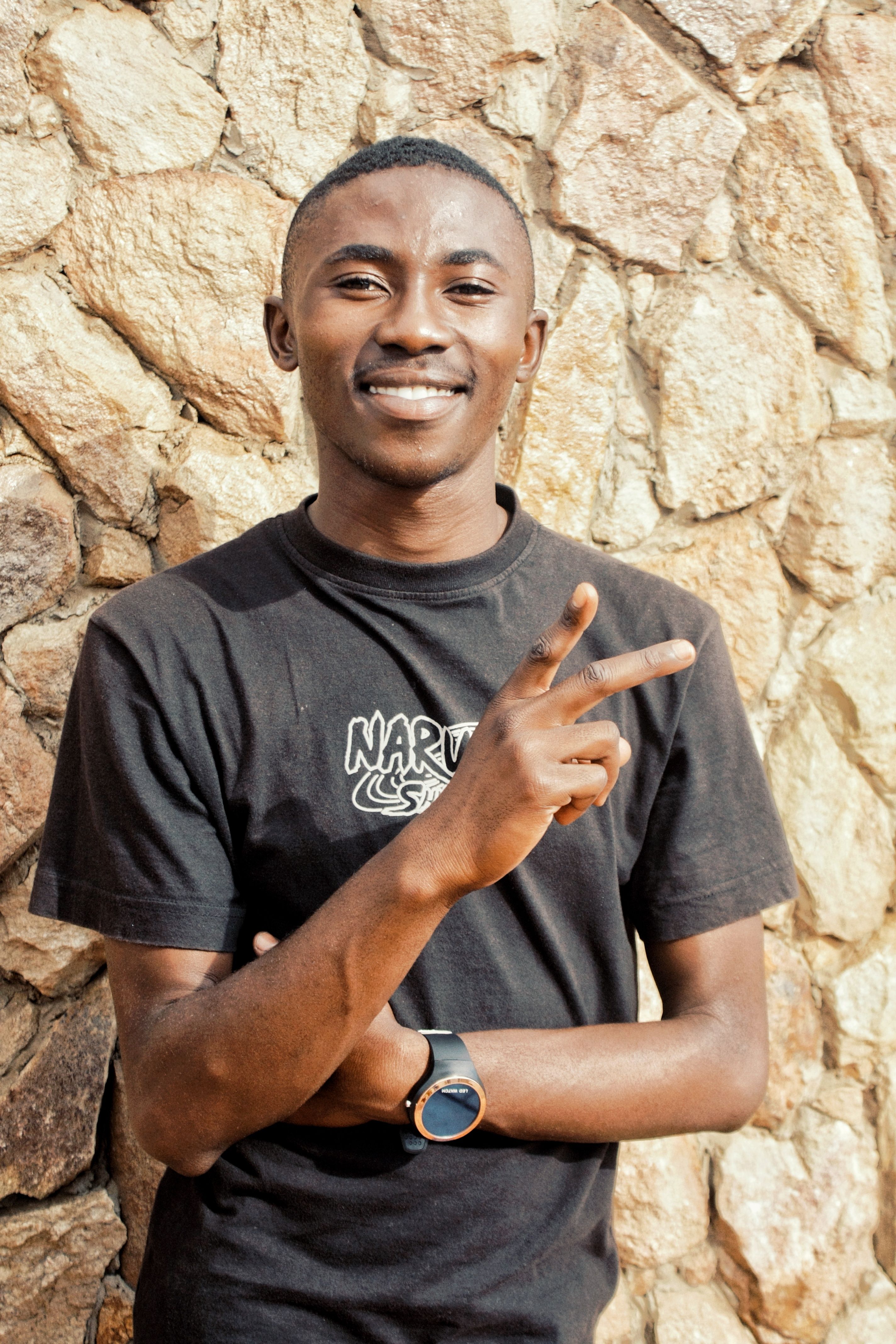 A smiling young man in a black T-shirt gestures peace against a stone wall.
