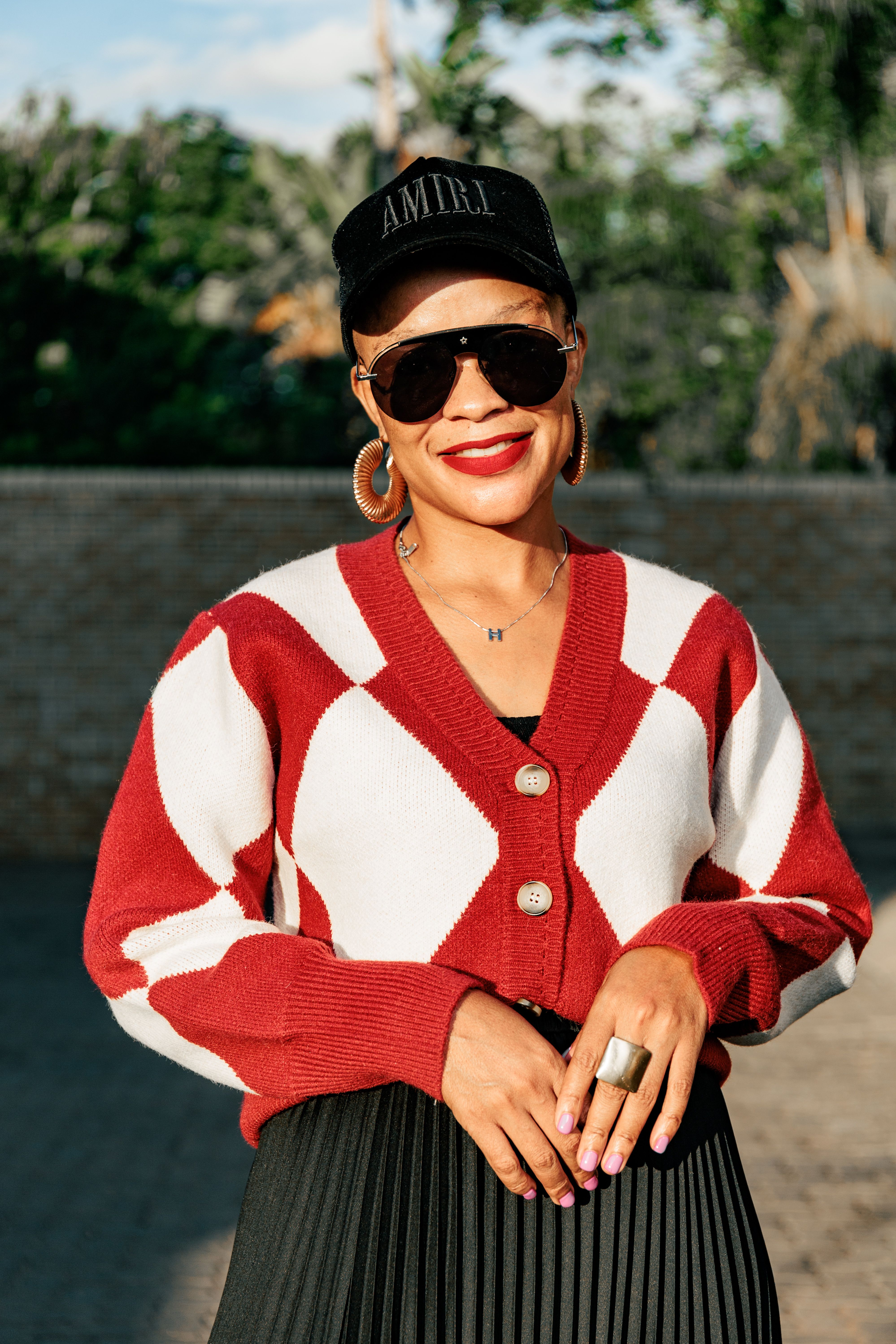 Stylish woman in sunglasses and cap smiling in a sunny outdoor setting, wearing a red and white sweater.
