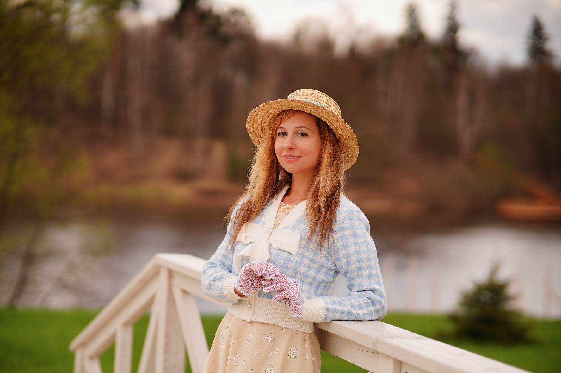 A stylish woman in a straw hat poses by a rural river landscape, embodying spring fashion.
