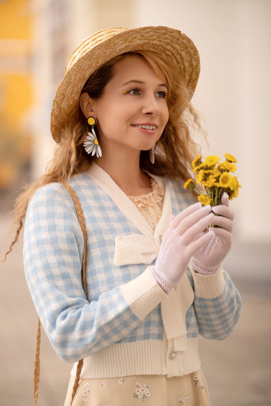 A cheerful woman in a vintage outfit smiling while holding a bouquet of yellow wildflowers outdoors.