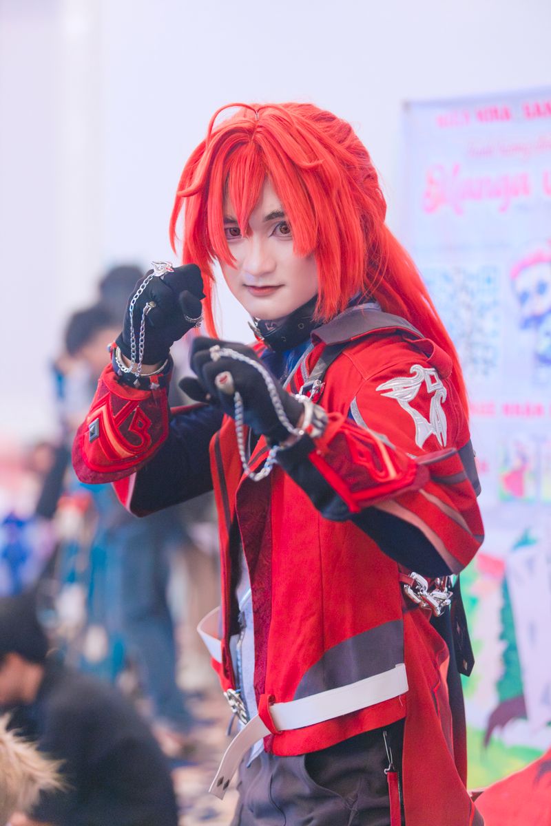 Vibrant cosplay character with red hair and outfit posing at a convention.