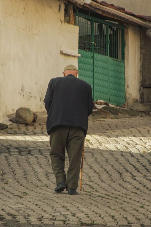 Free Elderly man using cane walking uphill on cobblestone street, illustrating determination and resilience. Stock Photo