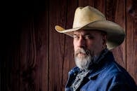 一位留着胡须、戴着帽子的粗犷牛仔，在质朴的木制背景前摆着姿势