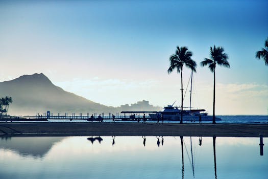 日落时分的夏威夷海滩宁静祥和，棕榈树、帆船与远山轮廓尽收眼底