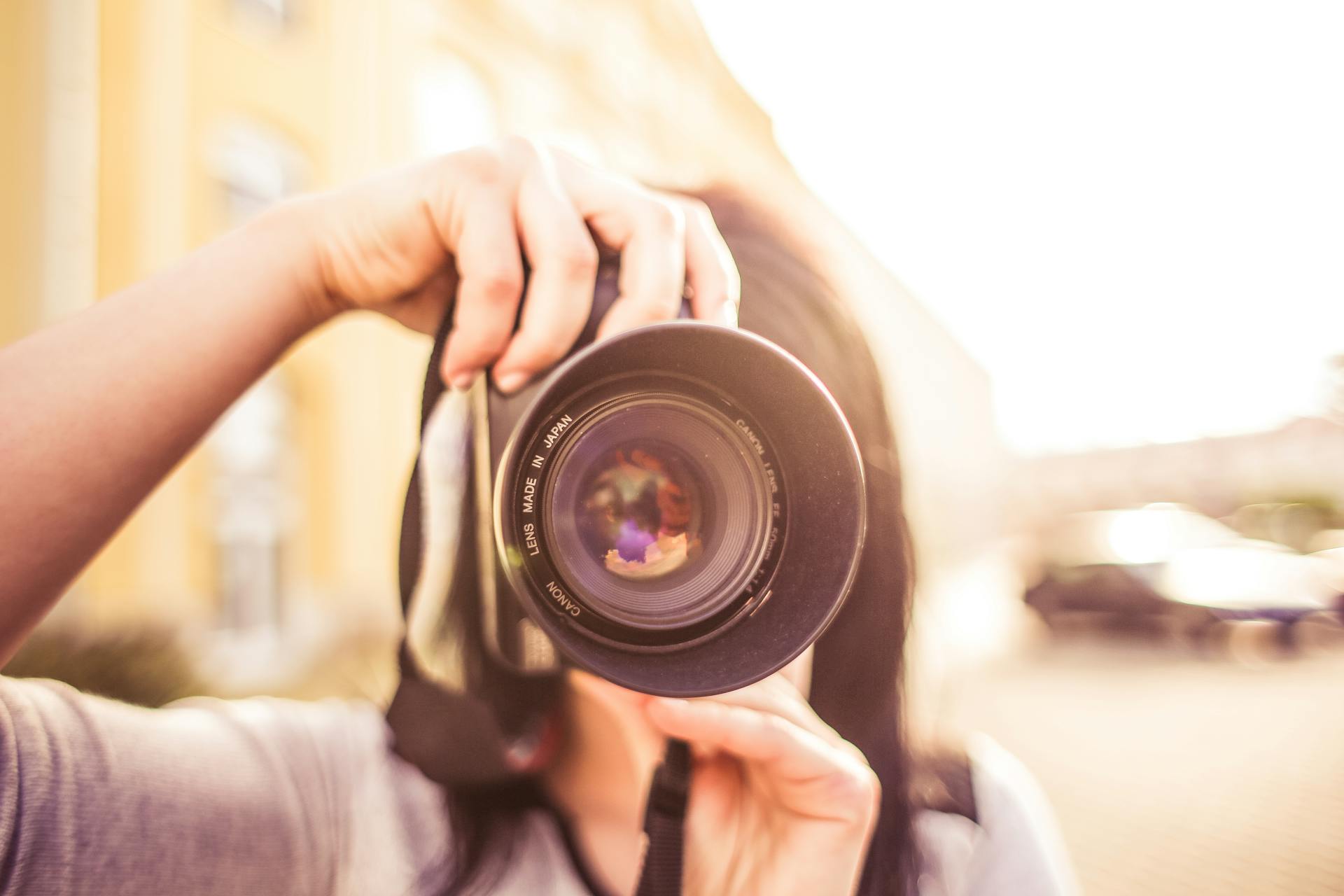 A young woman photographer focuses through a DSLR camera lens outdoors in bright daylight.