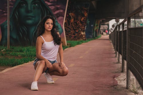 Photo of Woman in White Spaghetti-strap Tank Top and Denim Short Shorts Squat Posing by Sidewalk