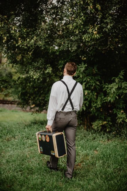 A man in suspenders walking through a grassy area carrying a vintage suitcase.