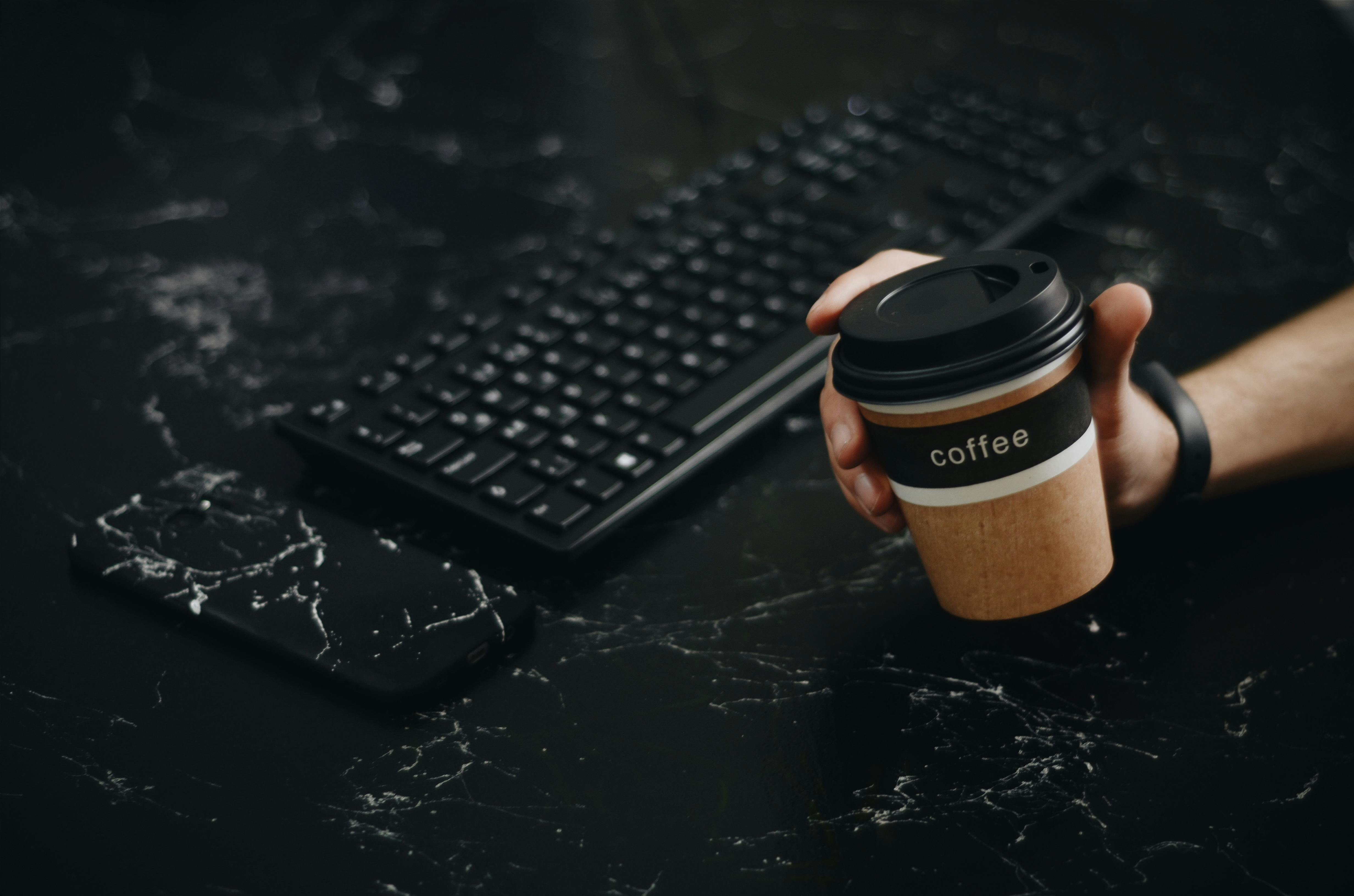 Person Holding Brown Disposable Cup Near Wireless Keyboard