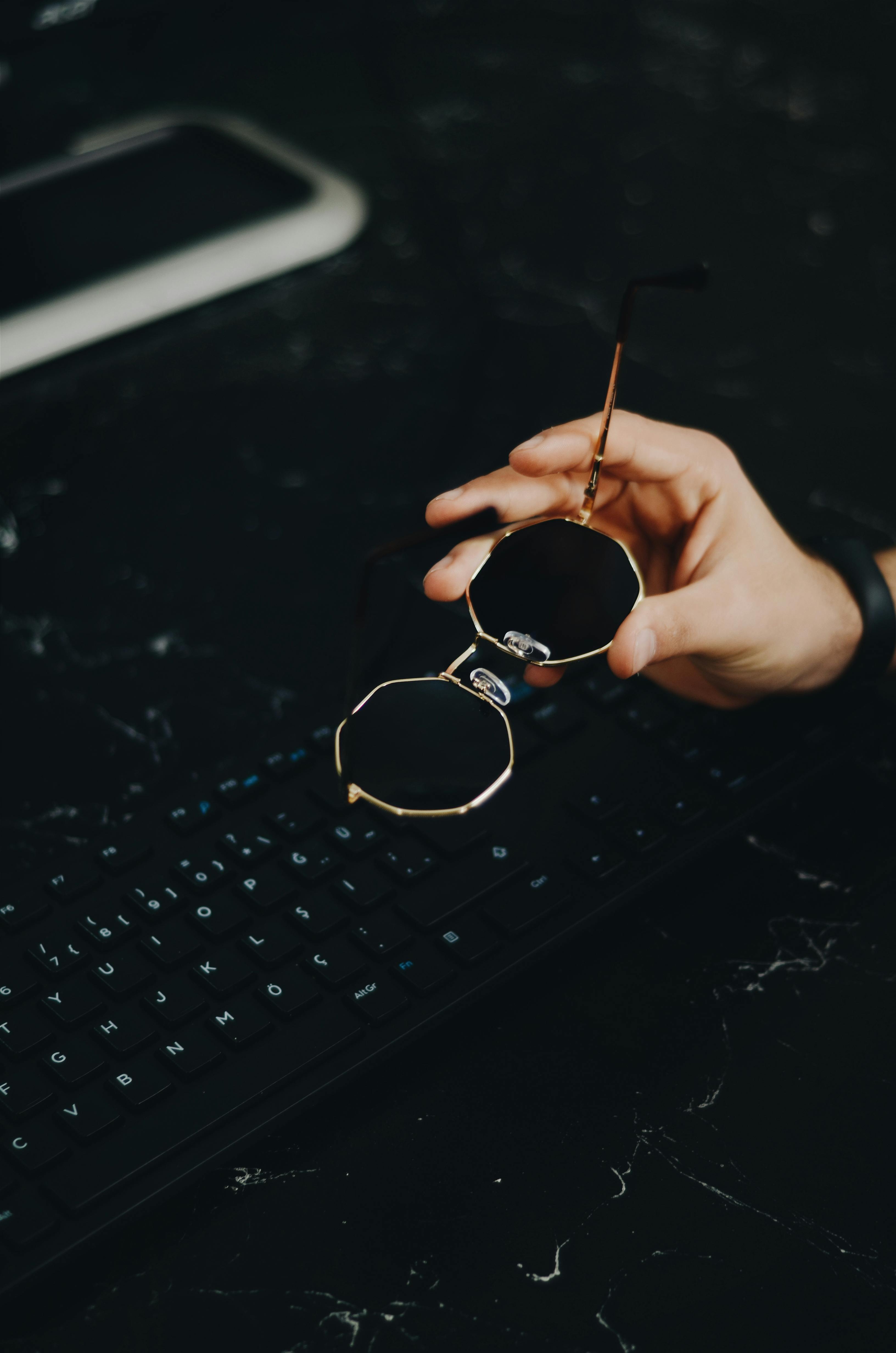 一只手拿着时尚的太阳镜，置于黑暗表面的黑色电脑键盘上方