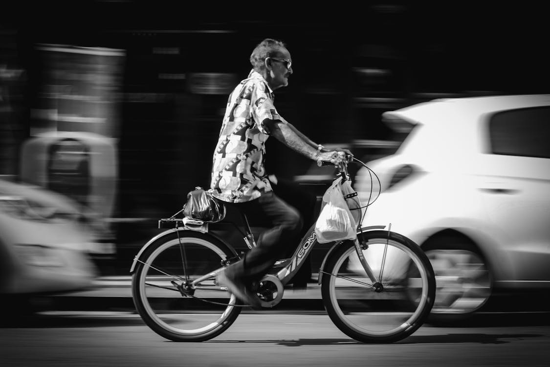 Greyscale Photo of Man Riding Bicycle on Road Passing Vehicles