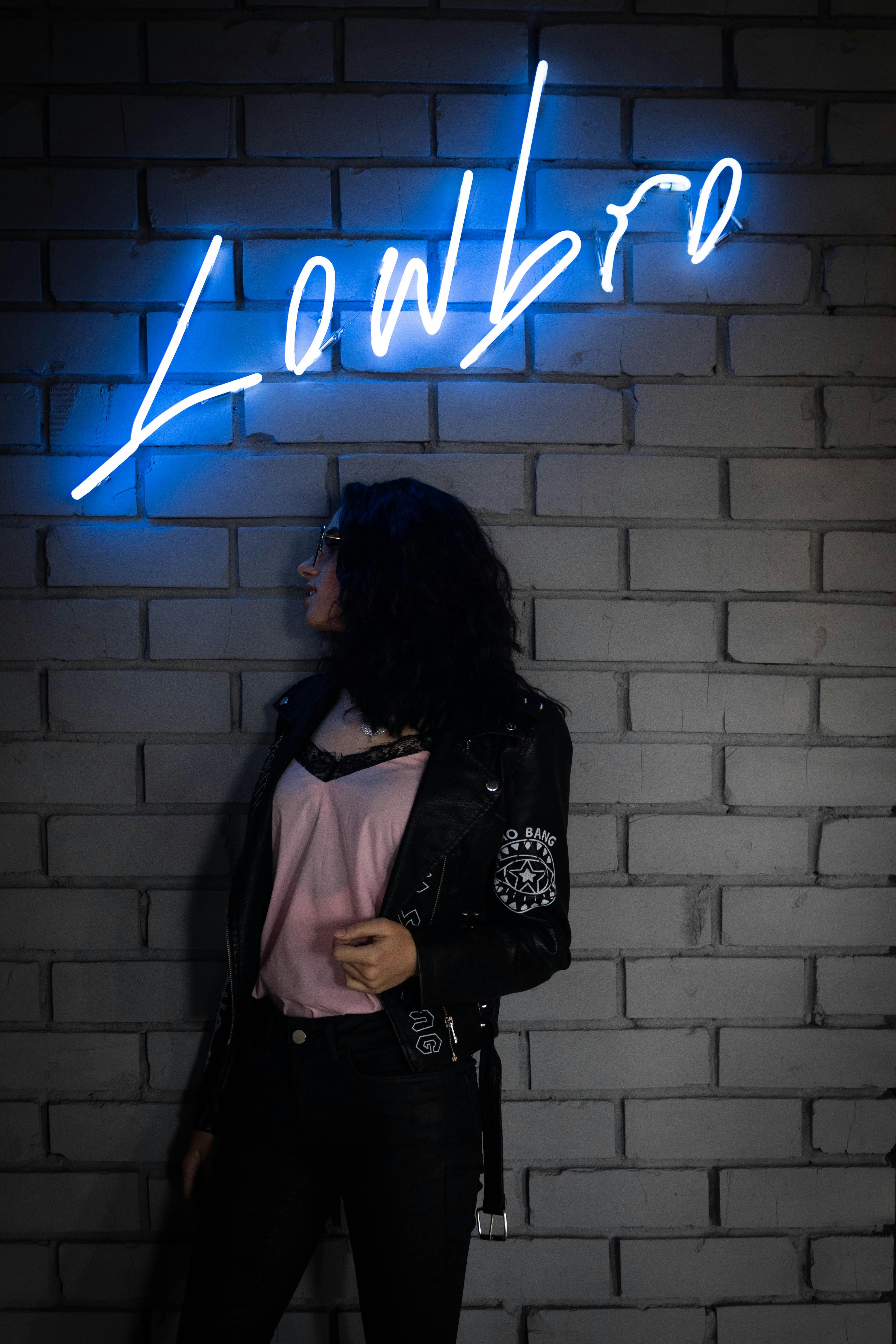 Trendy woman standing under blue neon Lowbrow sign against brick wall background.