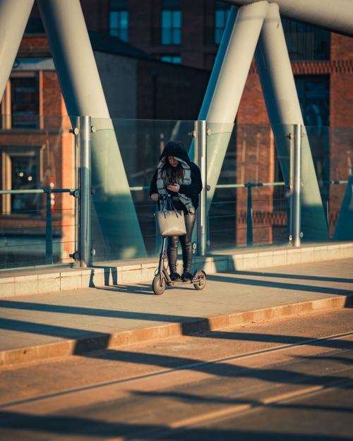 Free A woman on a scooter checks her smartphone on an urban bridge. Stock Photo