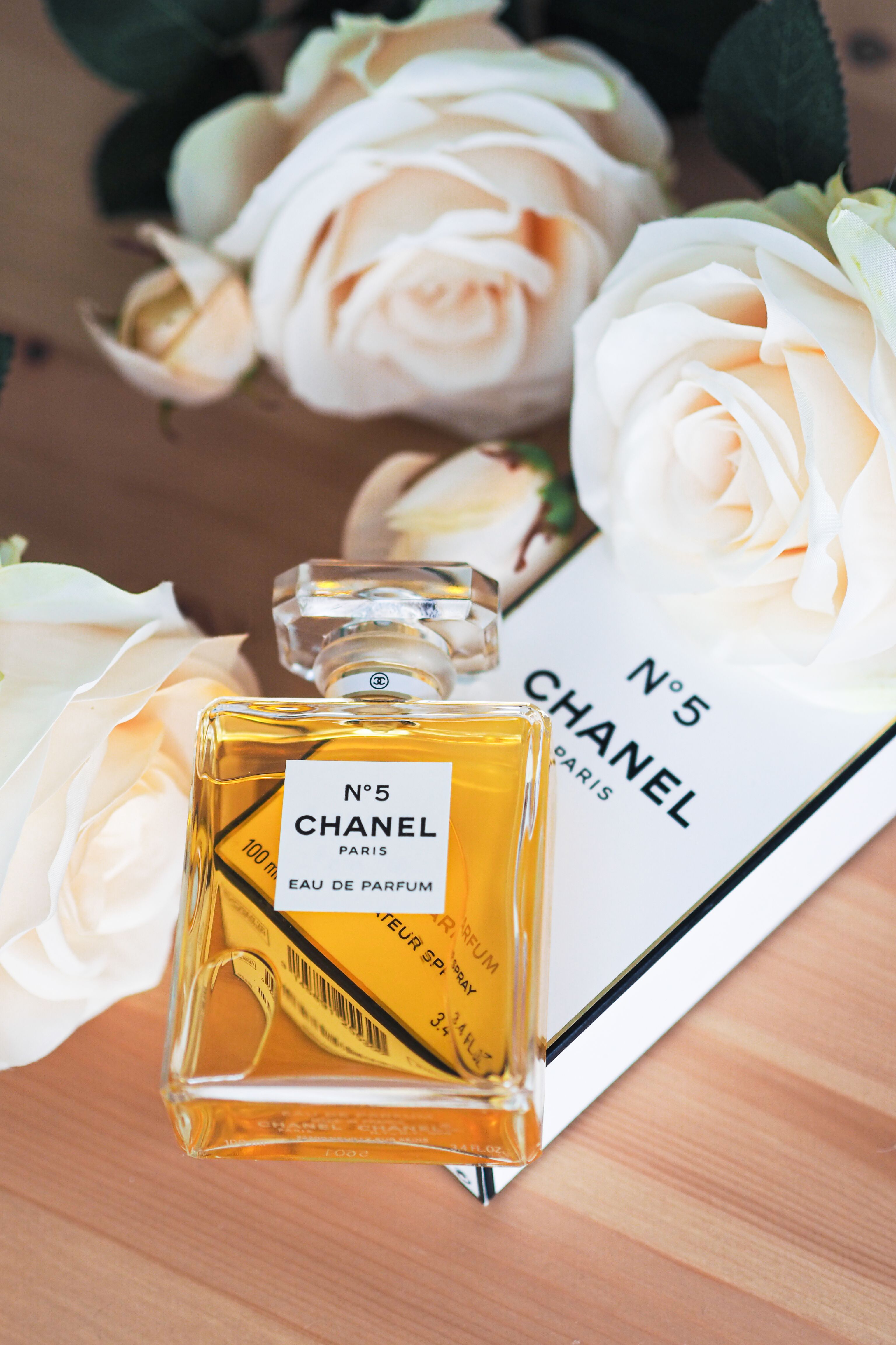 Chanel No. 5 perfume bottle surrounded by cream roses in a still life composition.