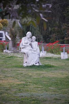 Elegant stone statue depicting a mother and child amidst lush greenery and vibrant flowers in a peaceful park setting.