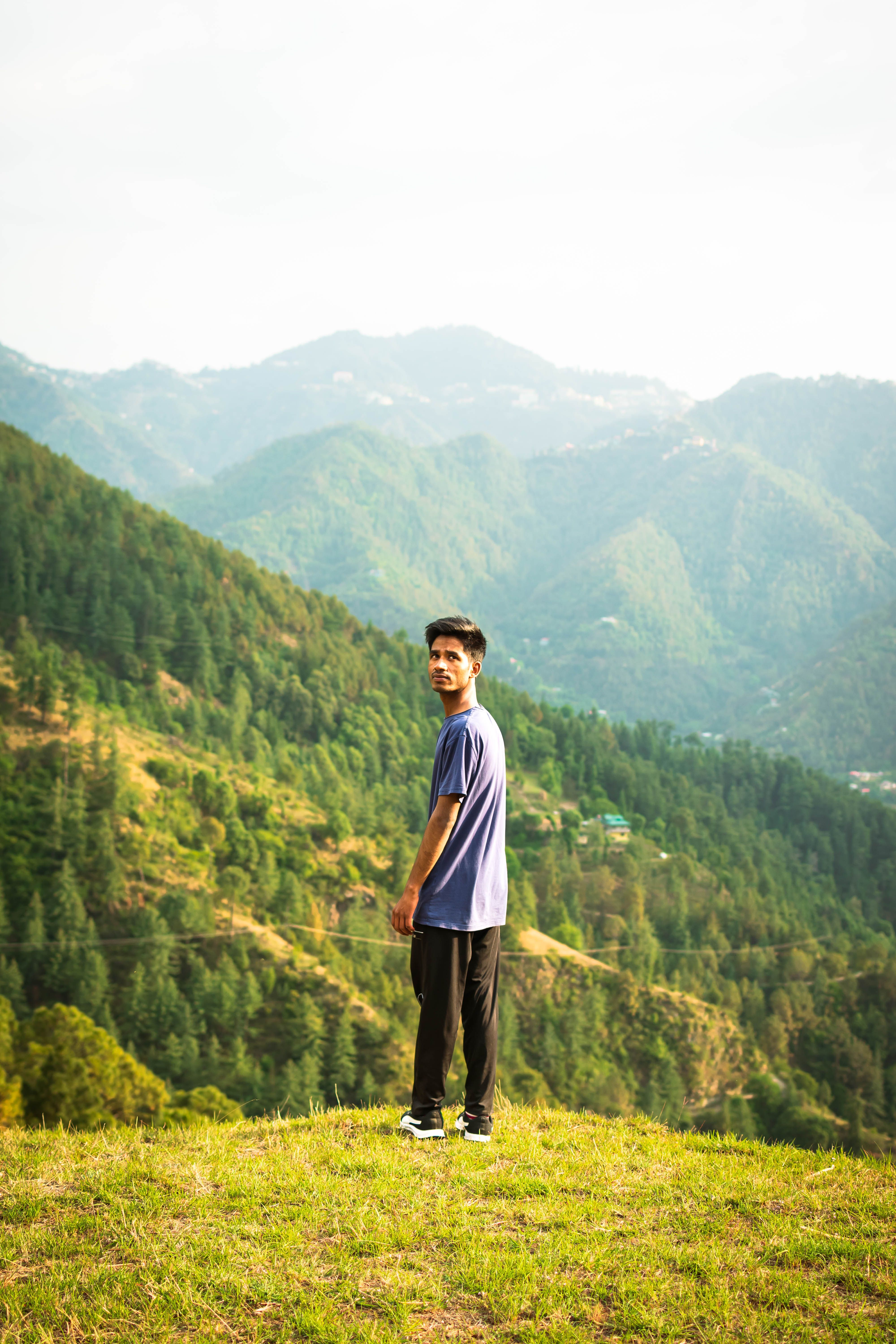 A young man stands on a grassy hilltop overlooking lush green mountains under a clear sky.
