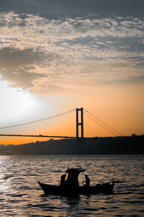 Free A fishing boat silhouette at sunset with a large bridge in the background, serene sea view. Stock Photo
