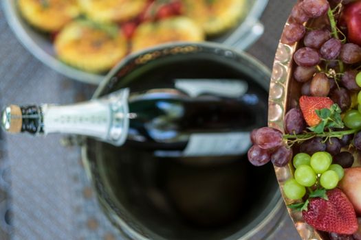 Overhead view of champagne bottle in ice bucket with fresh grapes and fruit platter on a sunny day.