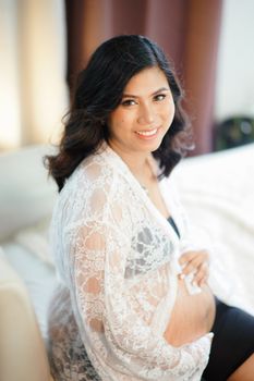 A cheerful pregnant woman sits on a bed, wearing a lace robe, capturing a moment of joy. - Fotografer Sidoarjo