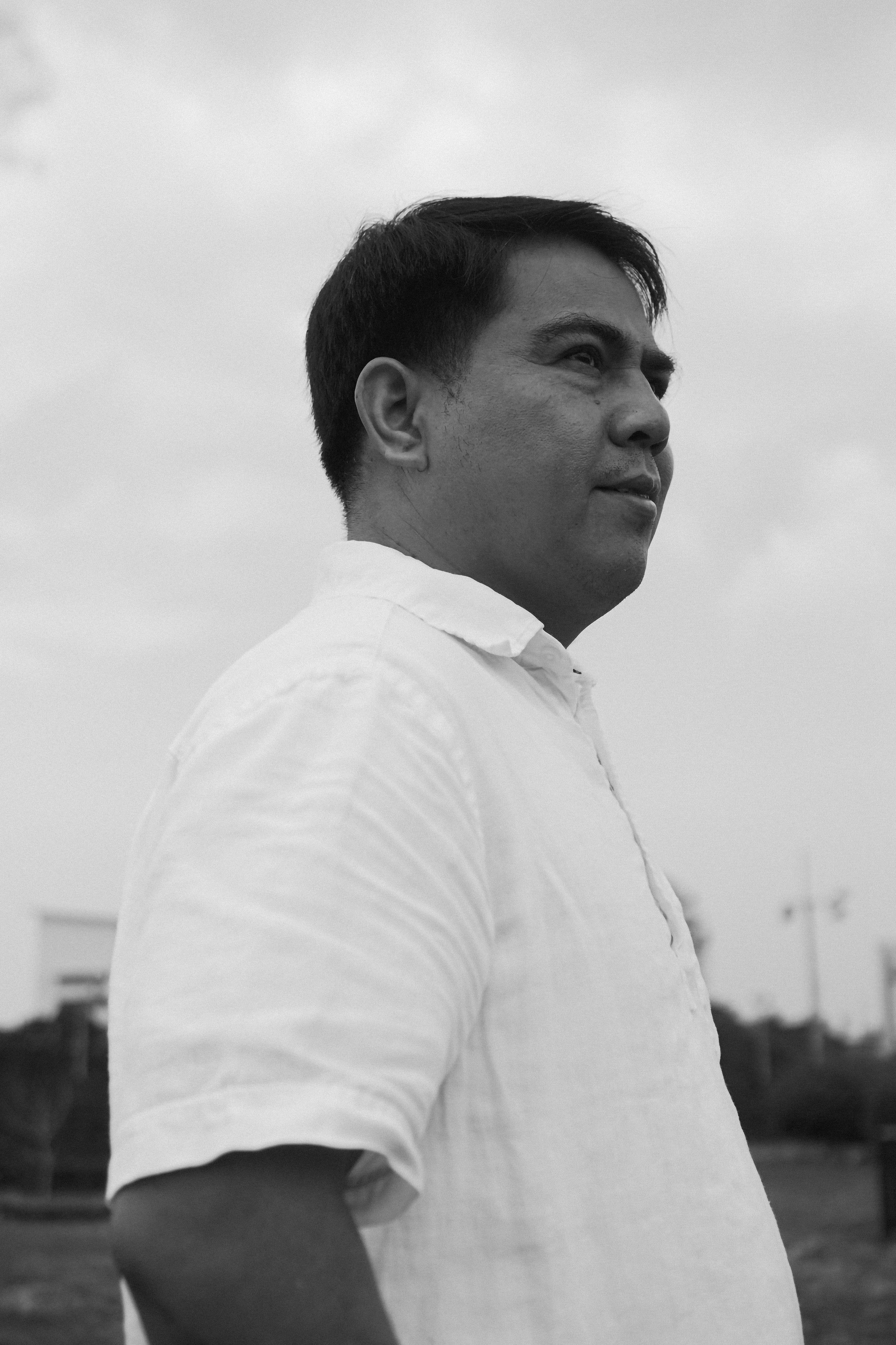Black and white photo of an Asian man in a white shirt outdoors, looking pensive.