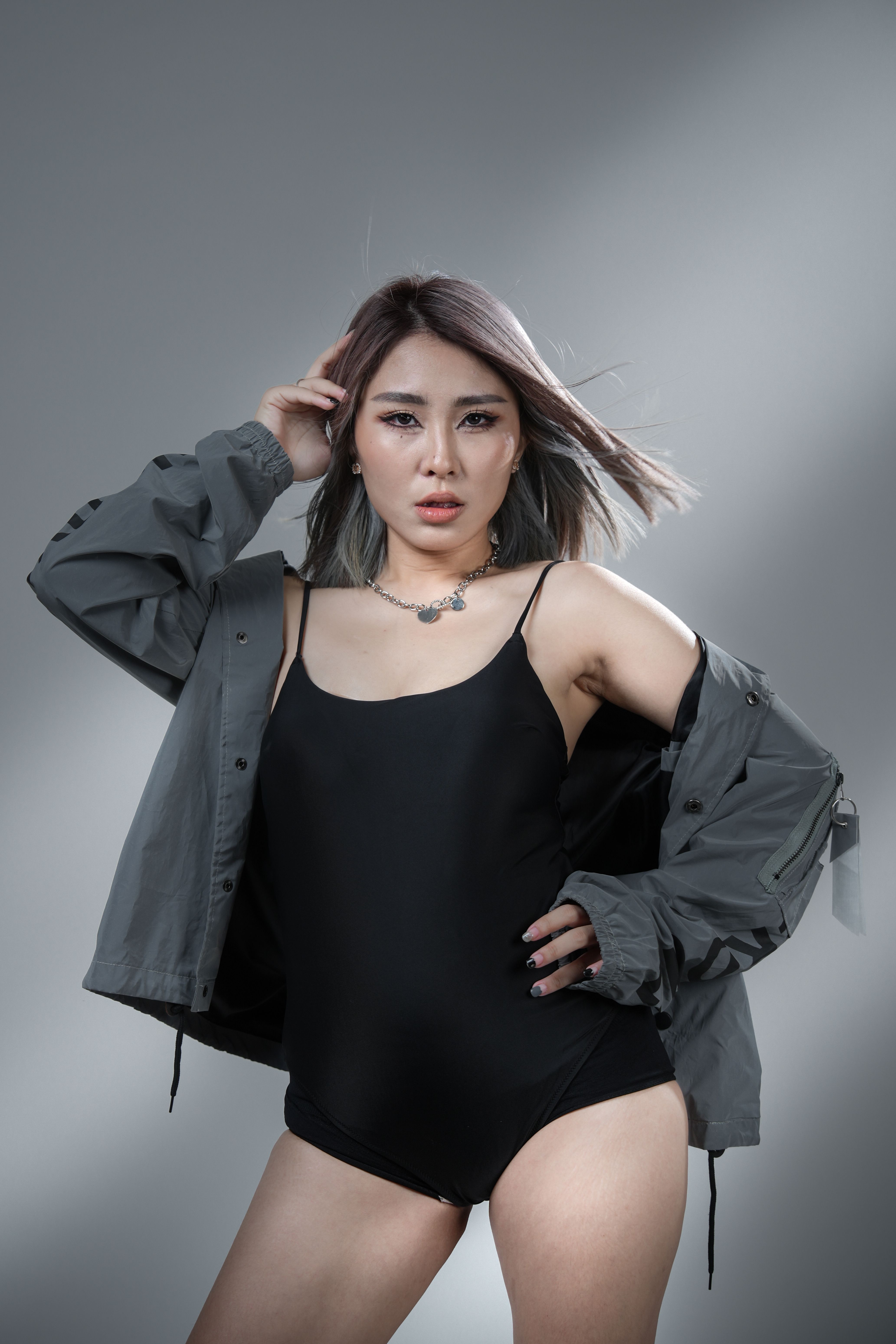 Asian woman poses confidently in a black bodysuit and gray jacket in a studio setting.