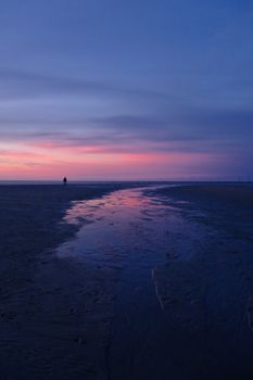 A lone person walks on a serene beach at dusk with a stunning sunset reflecting on the wet sand.