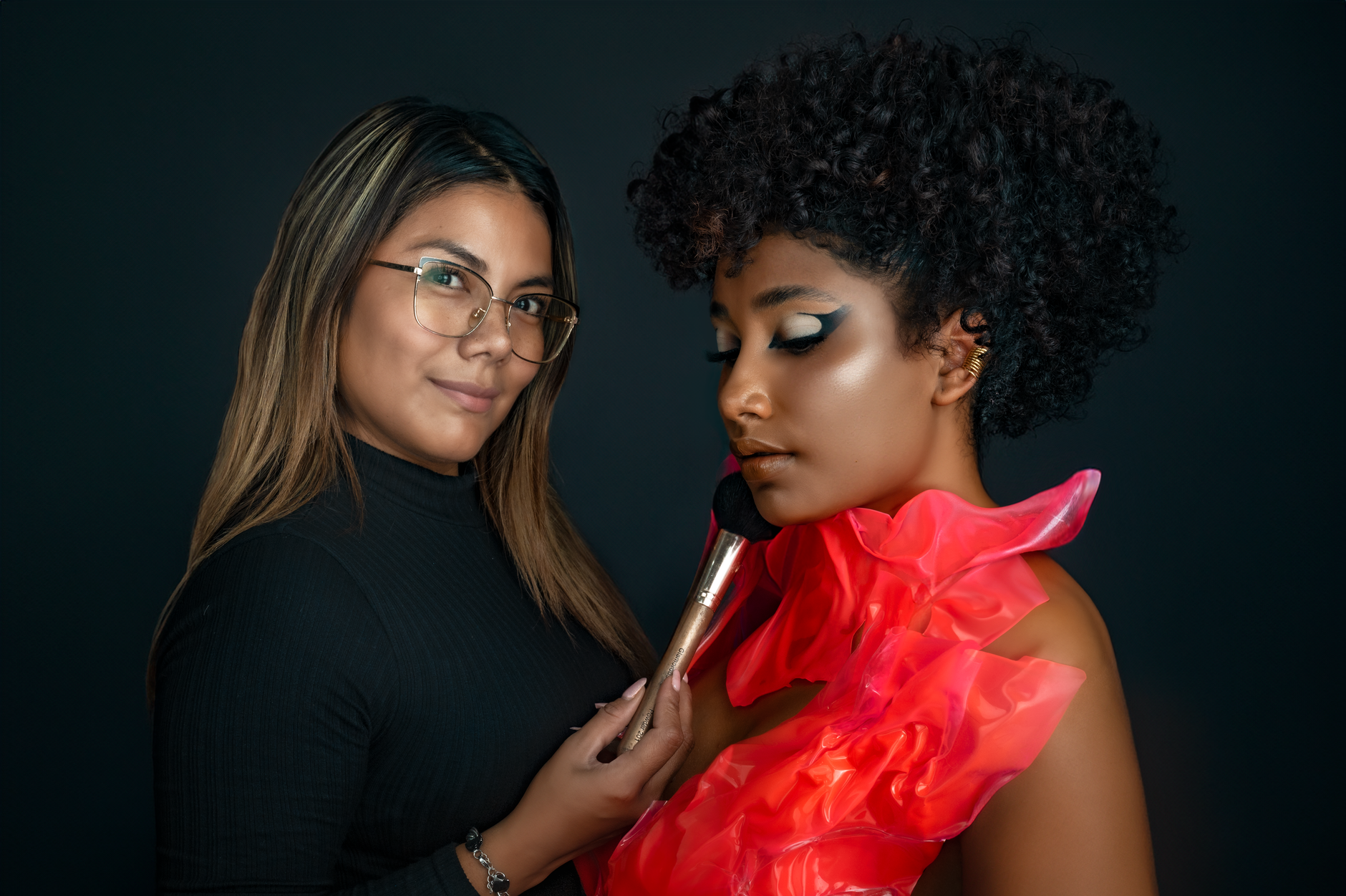 Two women in a studio, showcasing makeup artistry on a black background.