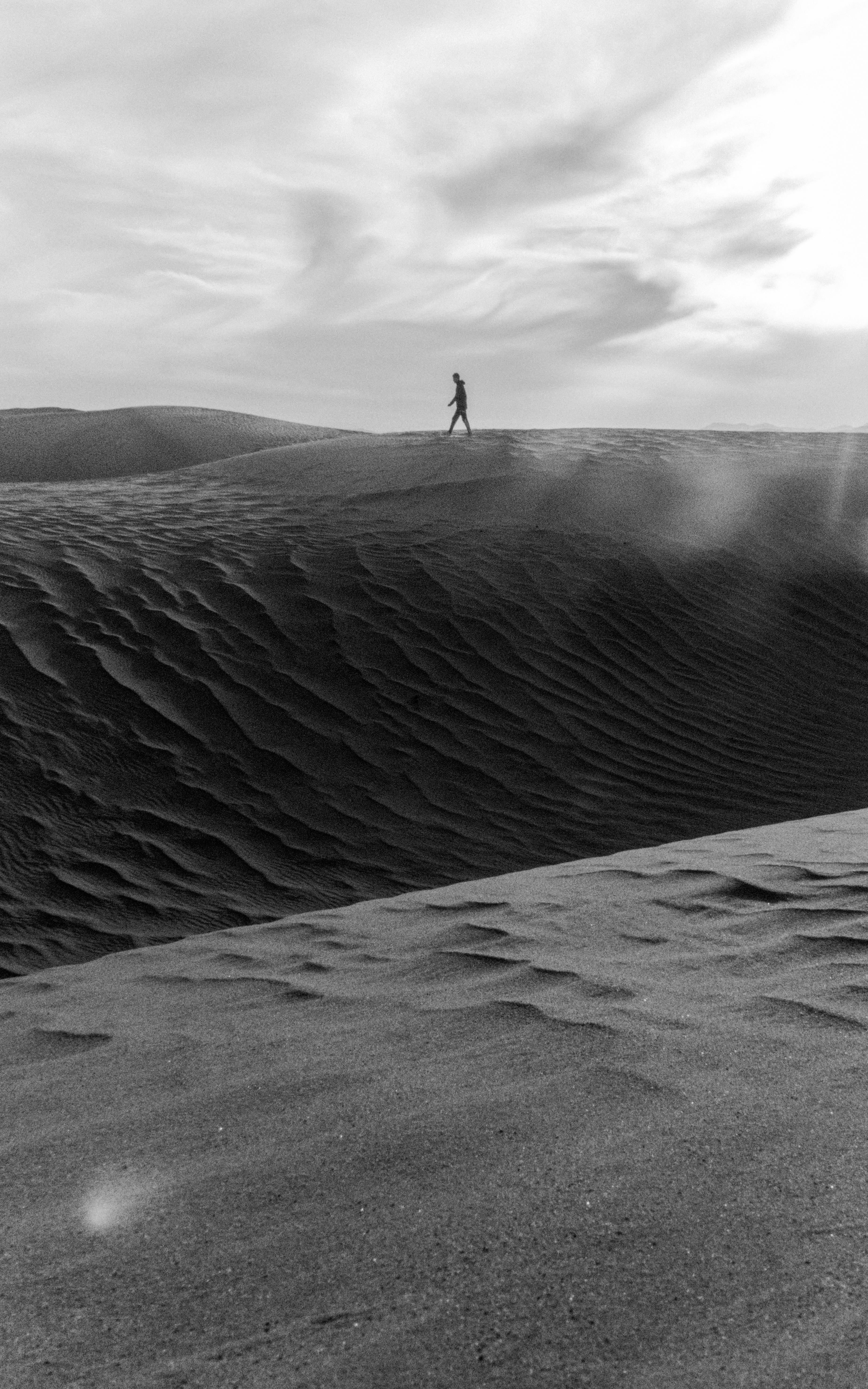 A solitary person walks over towering sand dunes in a dramatic black and white desert scene.