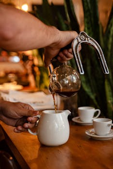 Close-up of a barista's hands pouring coffee into a ceramic pitcher in a cozy café setting.