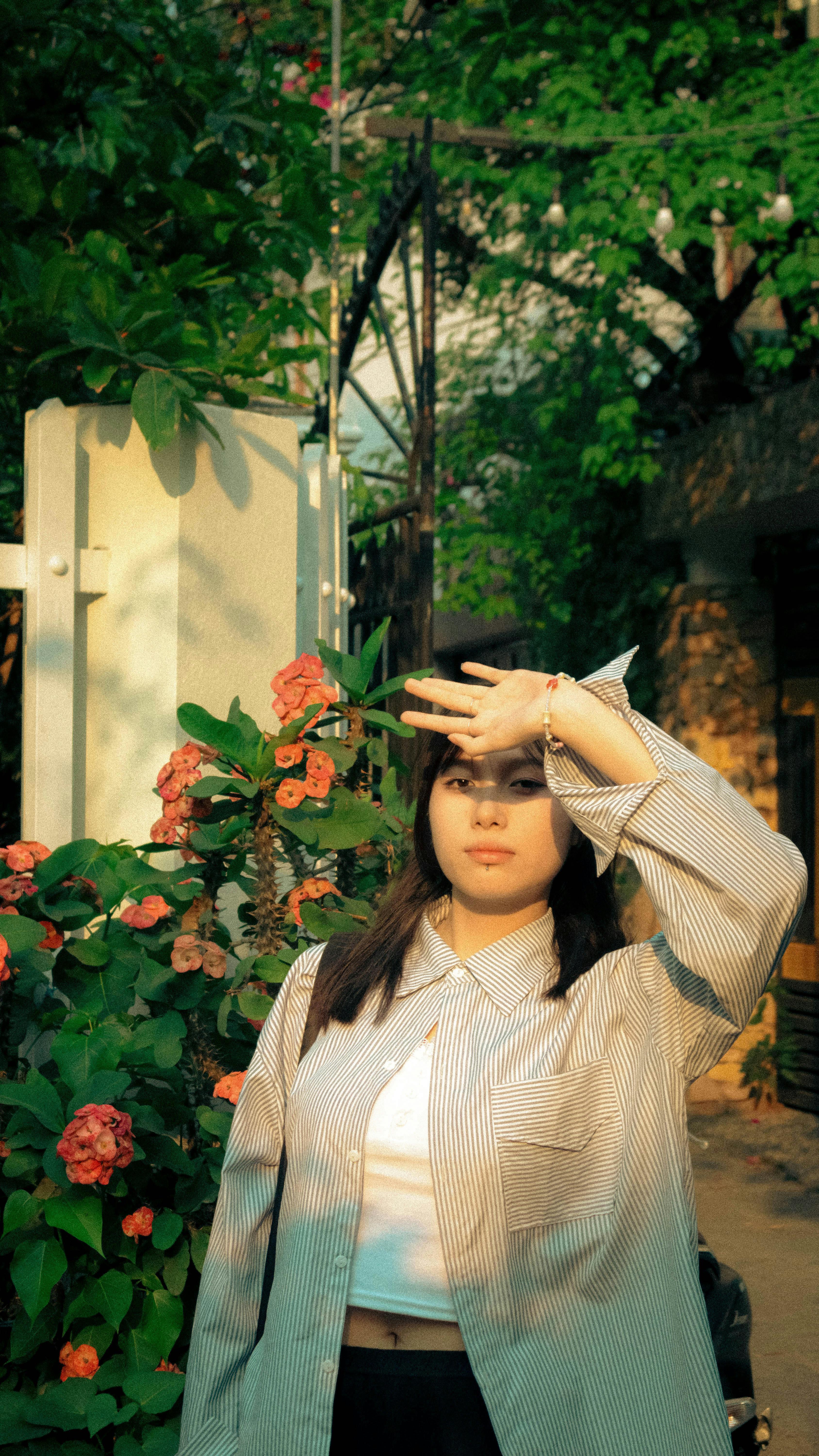 A young woman in a striped shirt shields her eyes from the sun amidst blooming flowers.