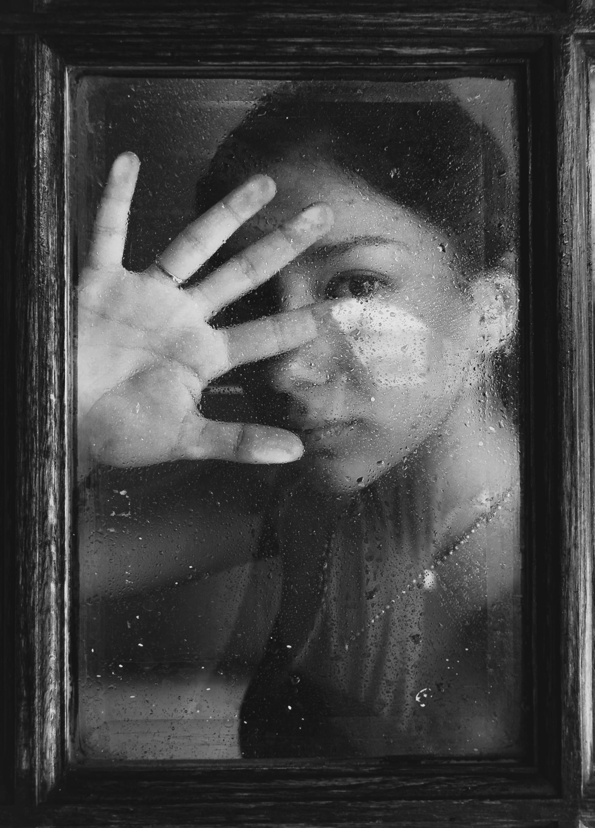 Black and white image of a woman touching a window with water droplets, creating an emotive scene.