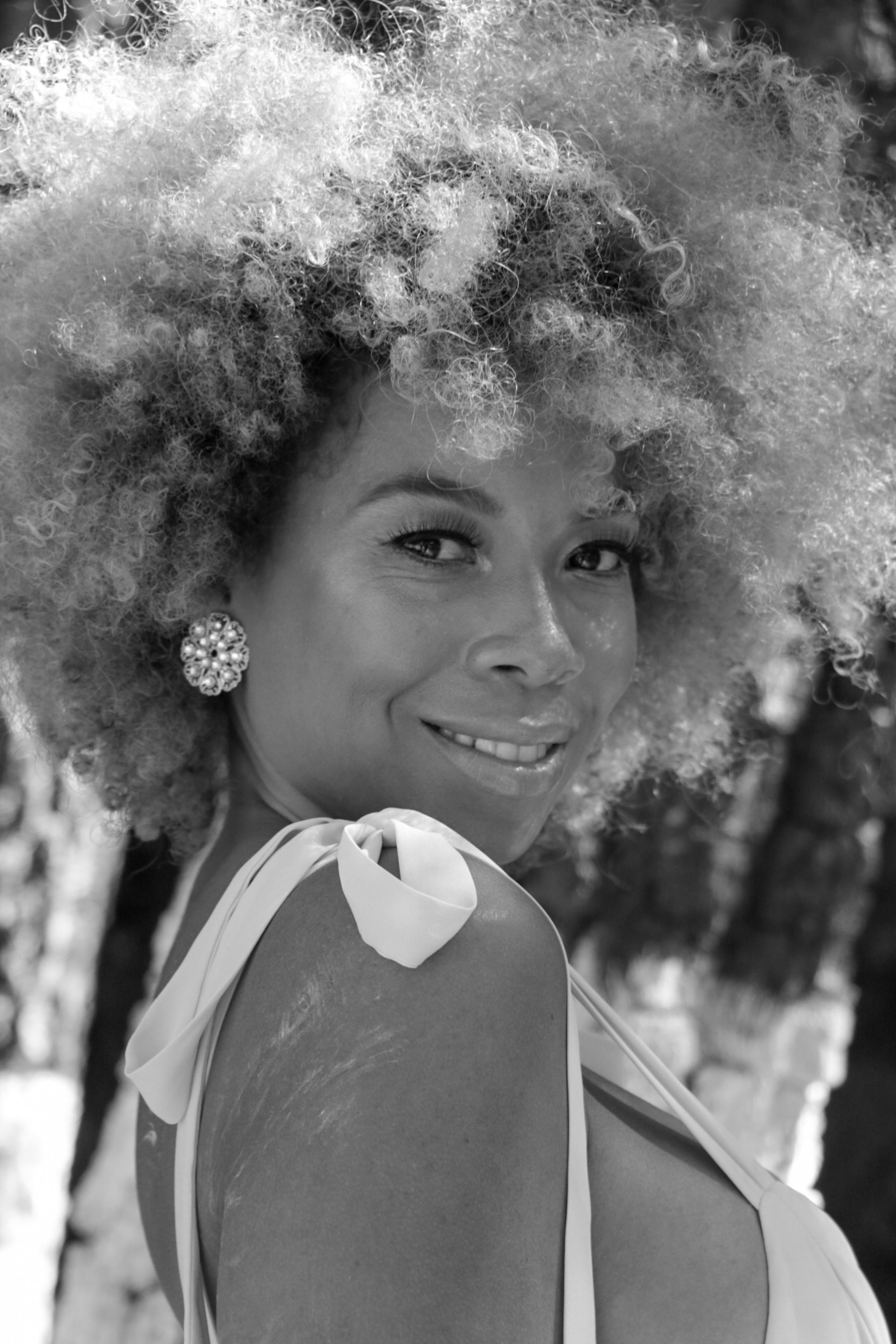 Black and white portrait of a smiling woman with afro hair and elegant earrings, set against a natural backdrop.