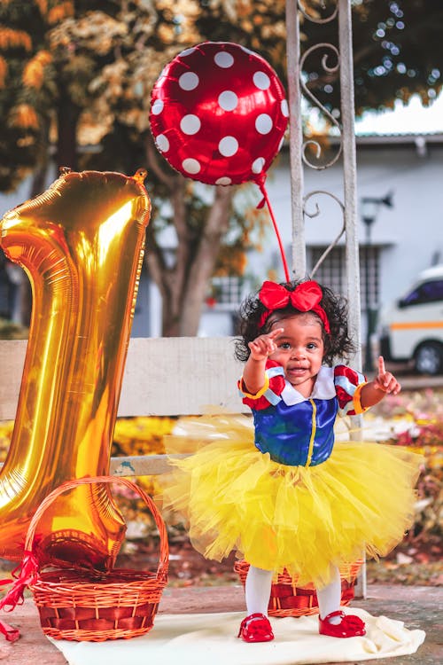 Photo of Baby Girl in Tutu Dress Standing Beside Large Gold Number 1 Balloon