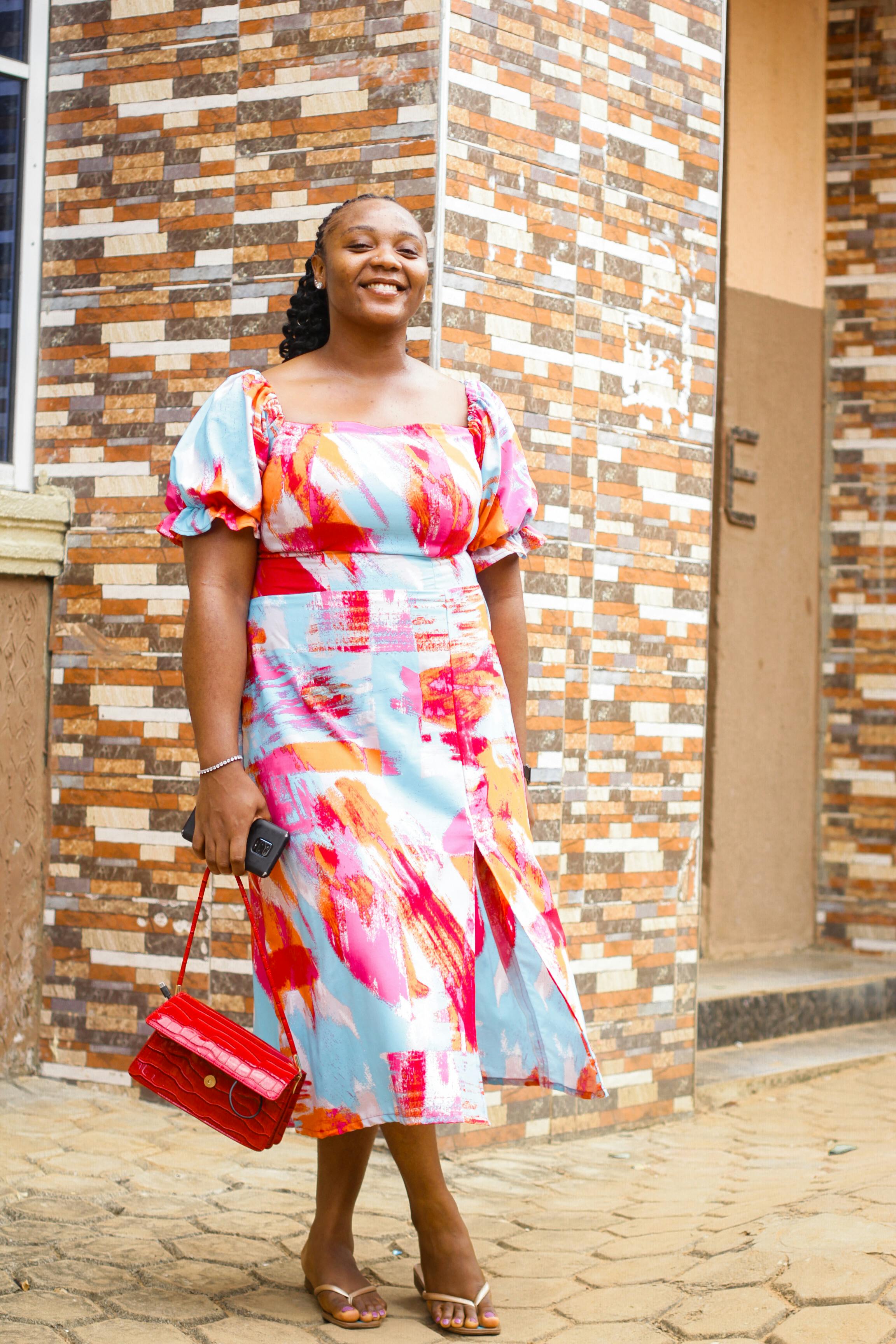 A woman wearing a vibrant, colorful dress smiles while standing outside a patterned building wall.