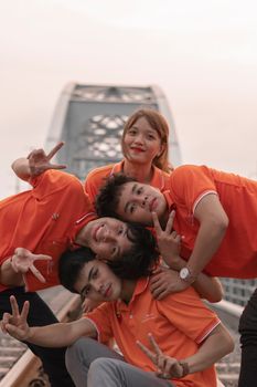 Five teenagers in orange shirts enjoy posing on a bridge, showing peace signs and smiling.