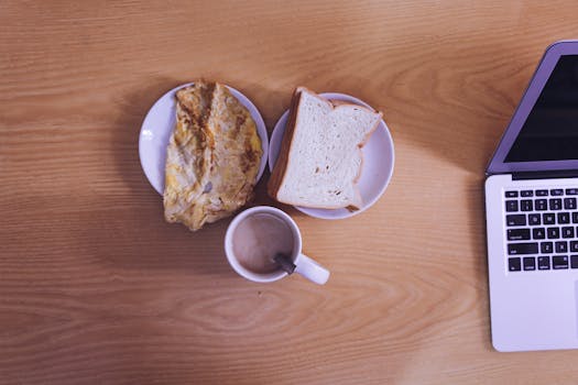 A cozy workspace setup with breakfast and laptop on a wooden desk.