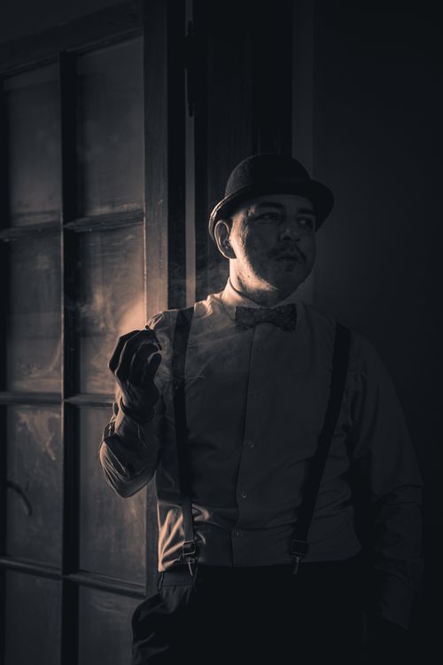 Free A vintage-style portrait of a man in a fedora and bow tie, smoking indoors with dramatic lighting. Stock Photo