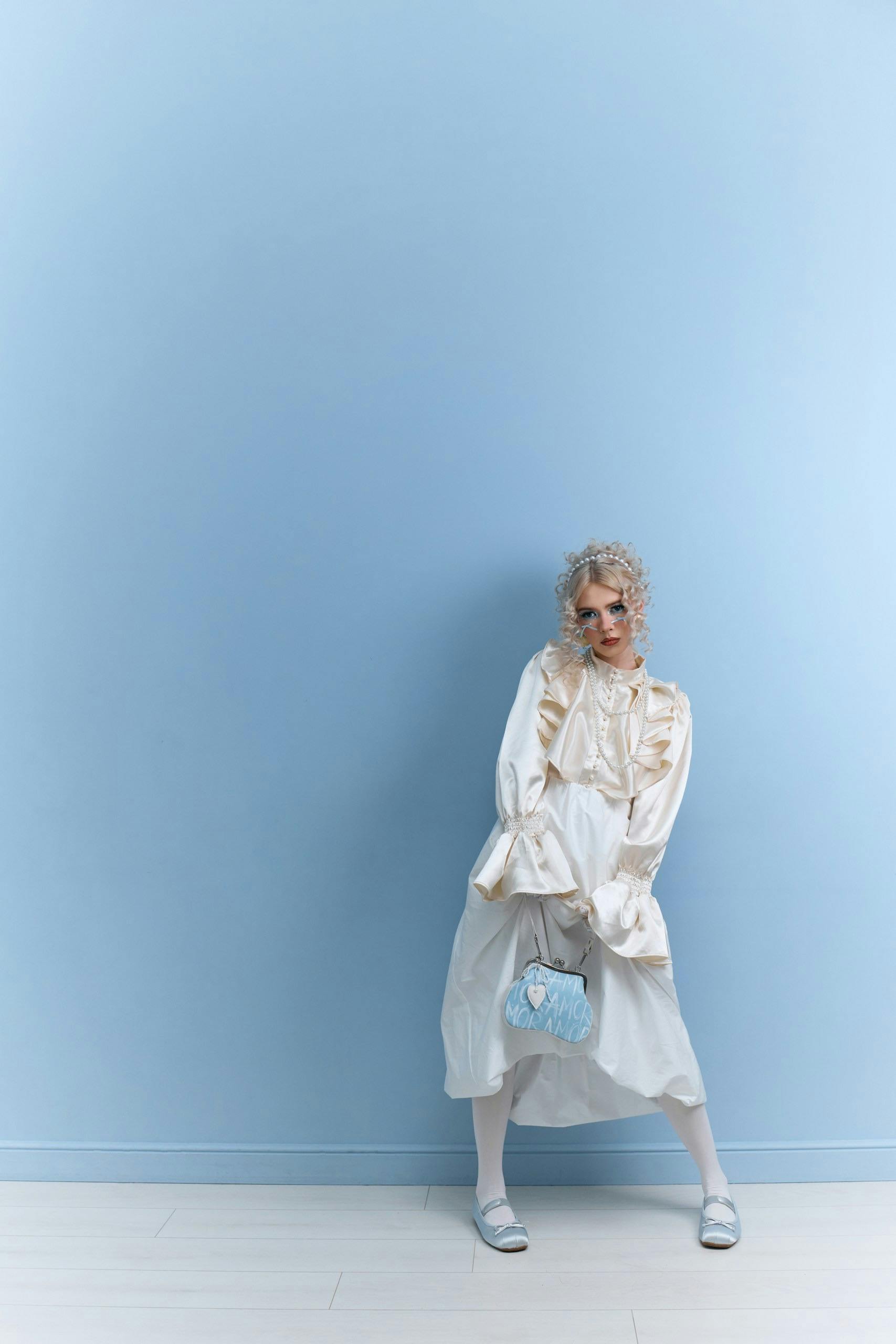 A stylish woman in a vintage dress poses against a solid blue background holding a blue handbag.