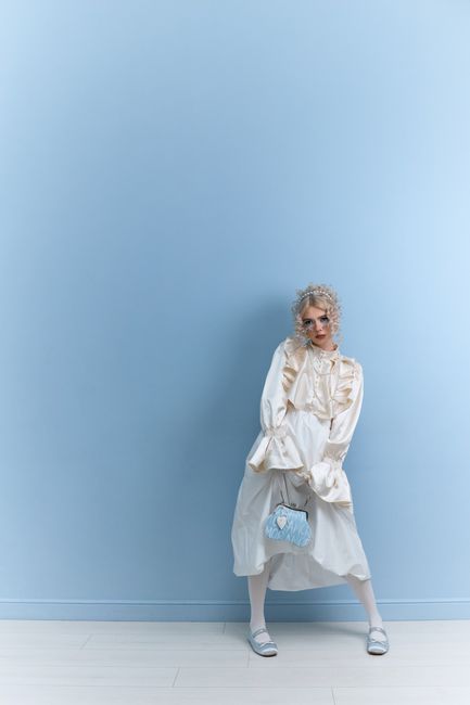 A stylish woman in a vintage dress poses against a solid blue background holding a blue handbag.