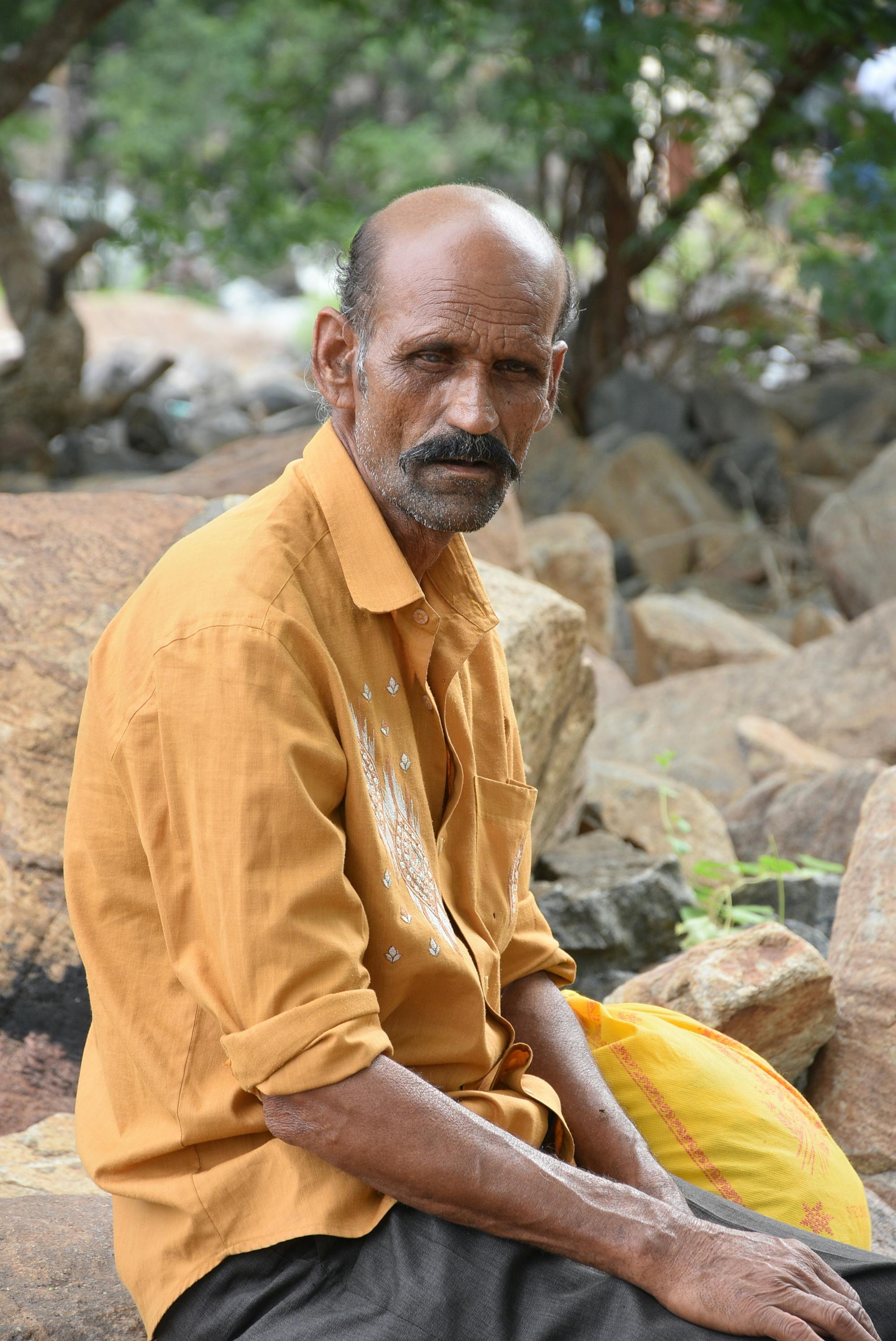Elderly man in a yellow shirt sitting on rocks in a park setting.