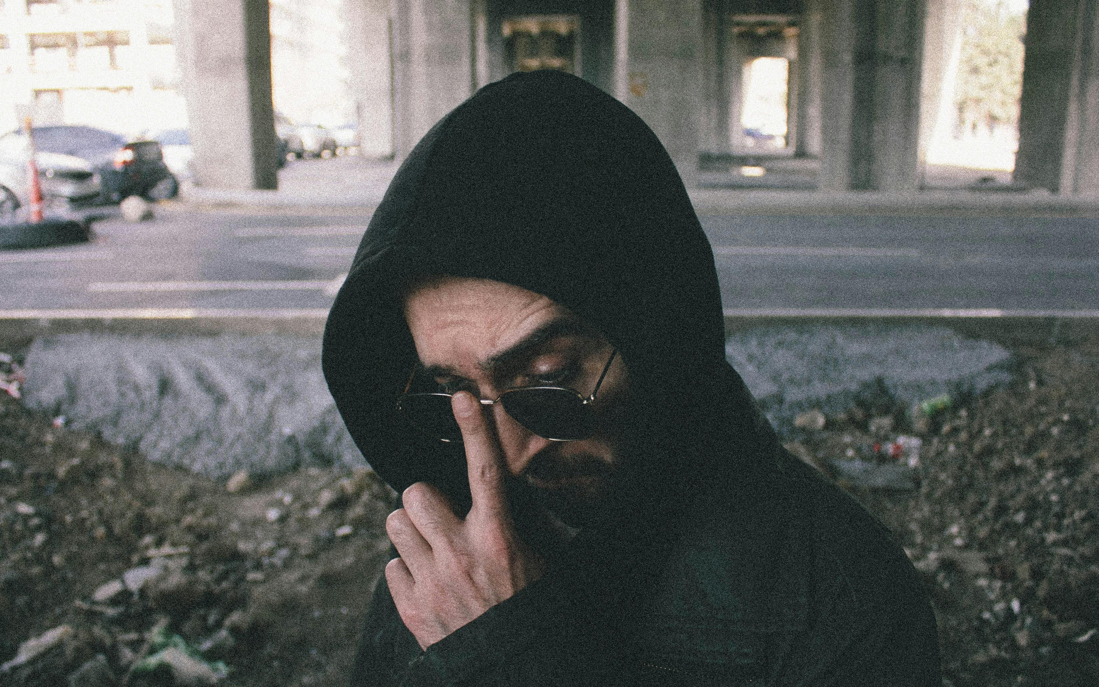 Street-style portrait of a man wearing sunglasses and a hood in an urban setting.