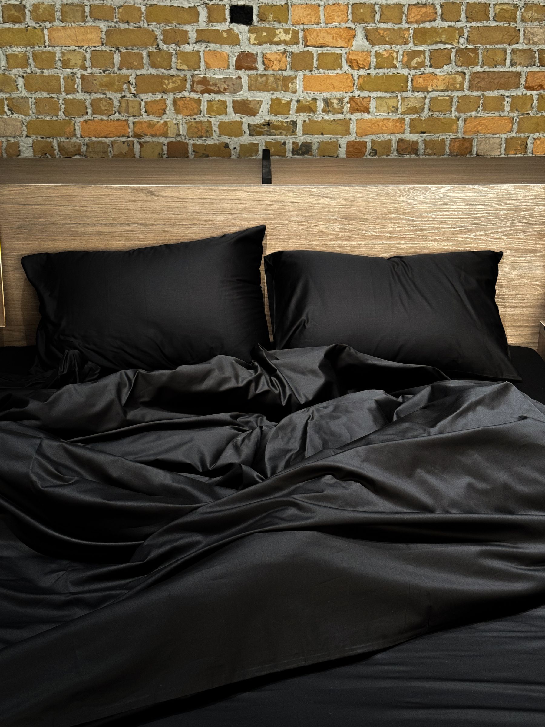 Bed dressed in black linen against a textured brick wall with warm timber accents