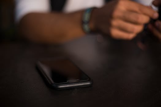 A smartphone rests on a table with a person holding an object in the blurred background, in a dark indoor setting.