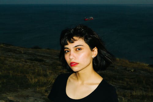 A woman with black hair standing on a hill with a boat in the background