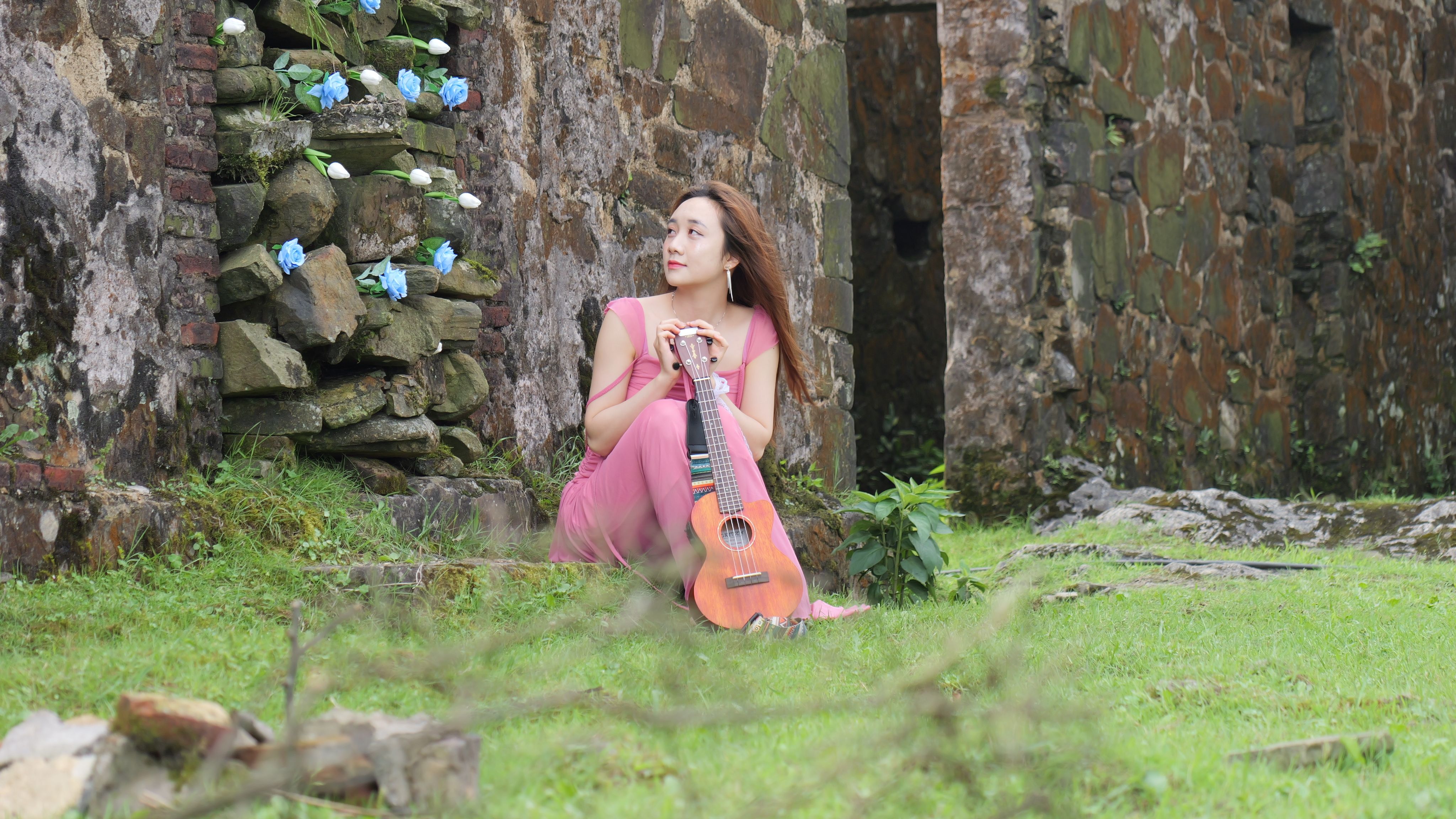 A woman in a pink dress sitting by a stone wall with flowers, holding a ukulele.
