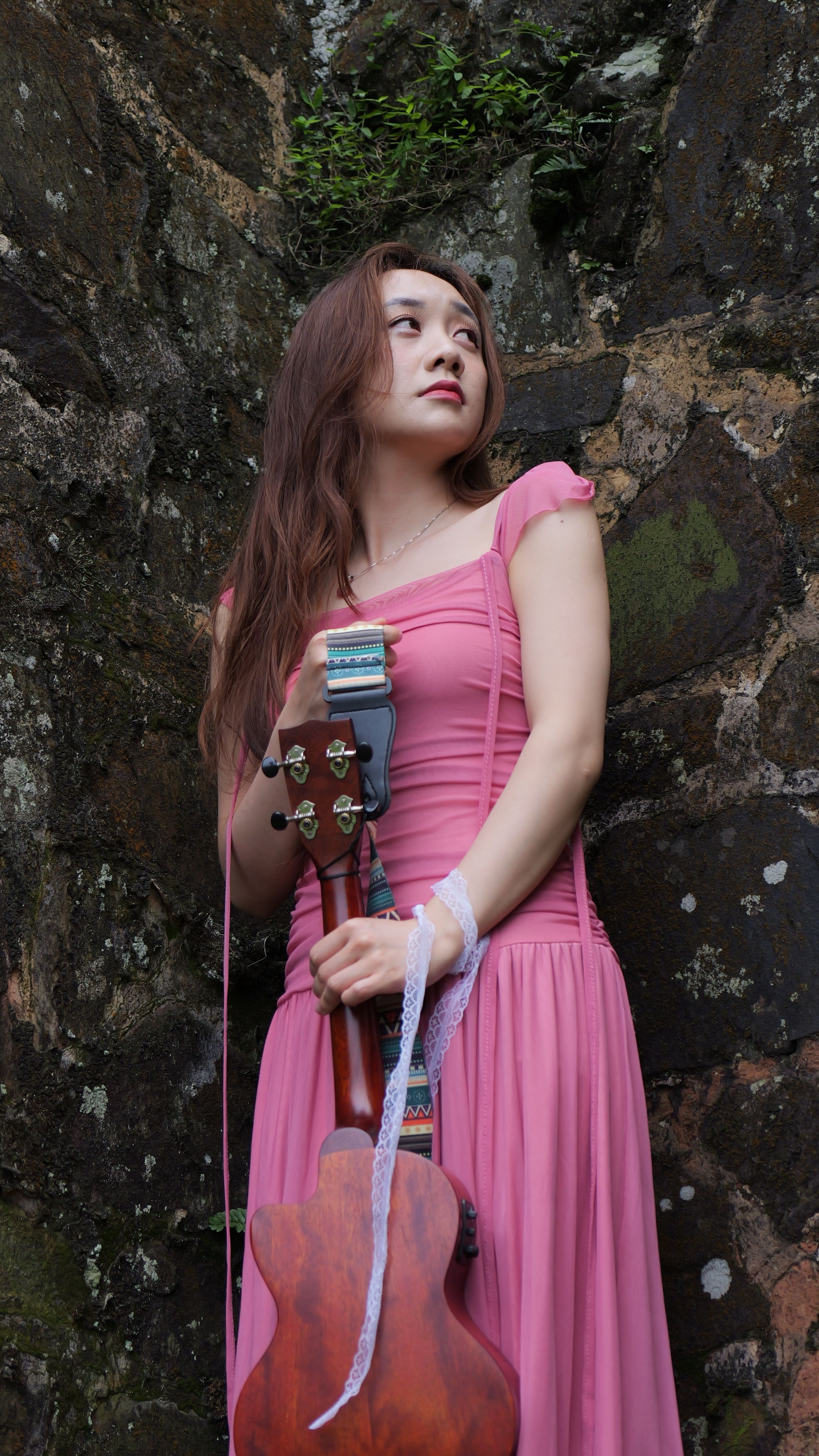 Young woman in a pink dress holding a guitar against a textured stone wall.