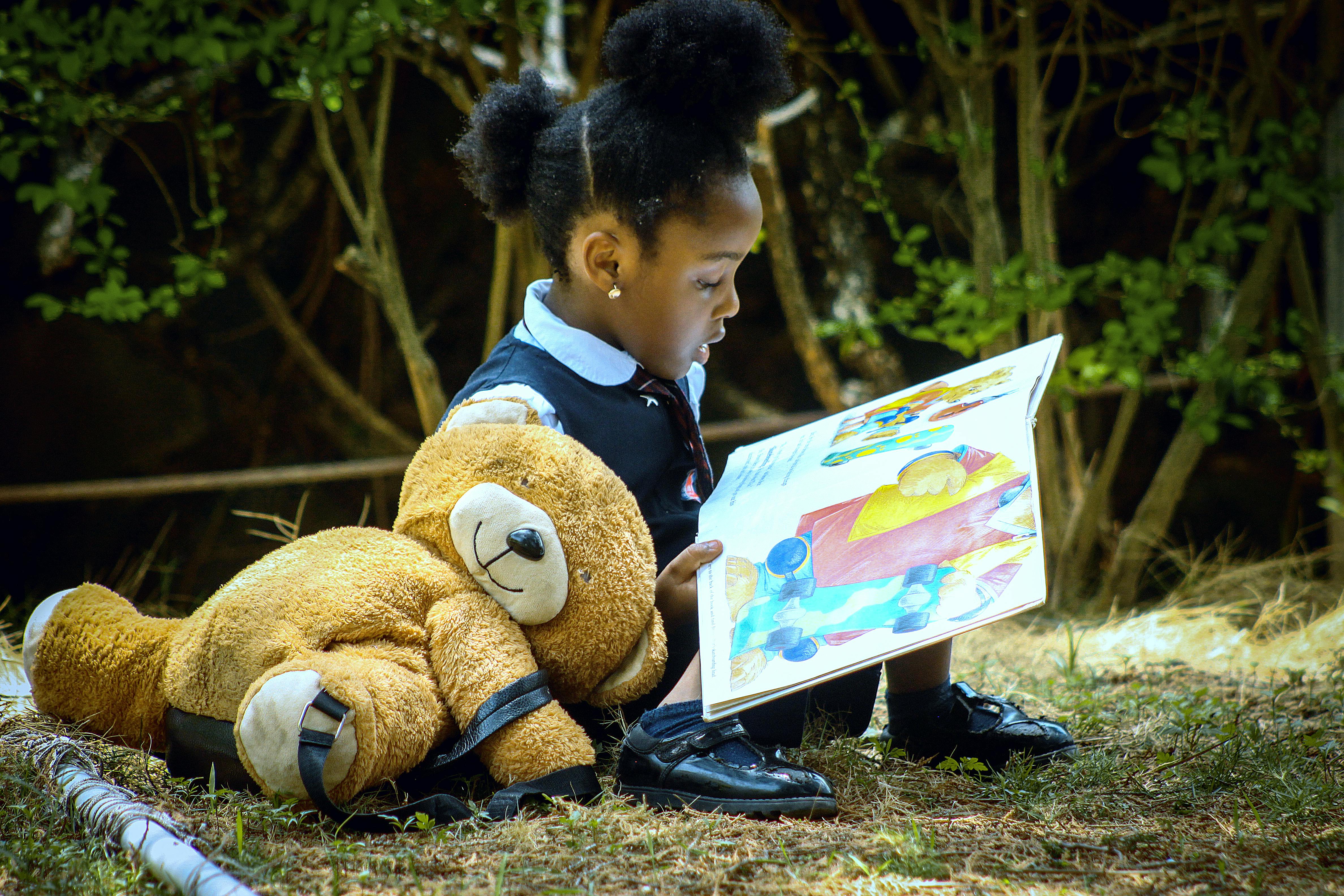 A young girl sits outdoors in a park reading a large colorful book with her teddy bear.